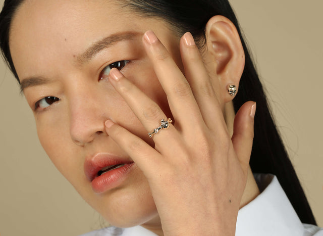 A women showing the side of her face with her hand in front of it. She is wearing very natural makeup. She is also wearing a yellow gold skull and bones ring and some skull stud earrings.